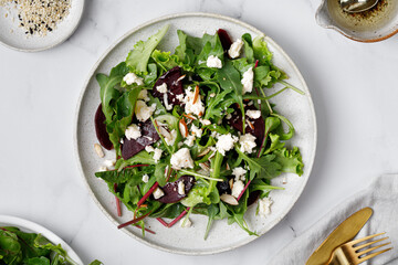 Salad with Beet or beetroot, green mix lettuce, nuts, feta cheese on white ceramic plate with fork, oil on marble white background, top view.