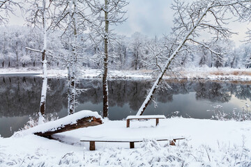 A snow-covered bench in a picturesque place by the river. Winter landscape by the river. The oaks after snowfall. The shore is reflected in the water of the river.
