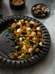 Salad with arugula, pumpkin, beetroot, ptitim, sesame, pistachios and feta on a black ceramic plate. Close up