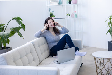 Woman sitting on a sofa with headphones and a computer in her apartment and listen to music and surfing the internet