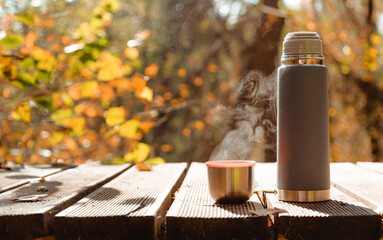 Thermos with mug of steaming tea on wooden table in autumn forest.