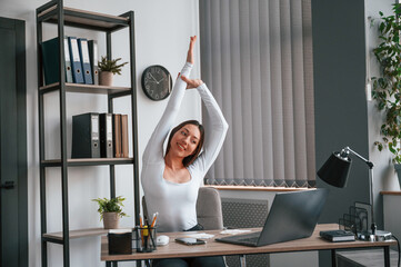 Takes a break, stretches her hands. Young beautiful woman in white clothes is in the office