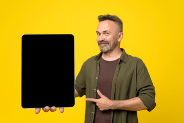 Happy smiled stubble and mustached mature man in green shirt pointing to his tablet pc with black...