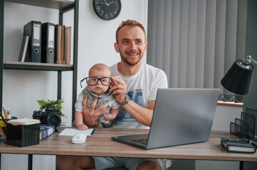 Big glasses. Sitting by the table with laptop. Father with his newborn son is indoors at home