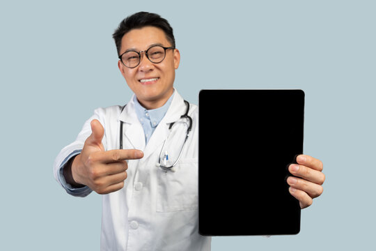 Satisfied Mature Chinese Male Therapist In White Coat, Glasses Pointing On Tablet With Empty Screen