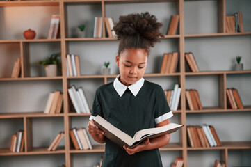 Standing and reading the book. Cute african american girl in school uniform is at home library