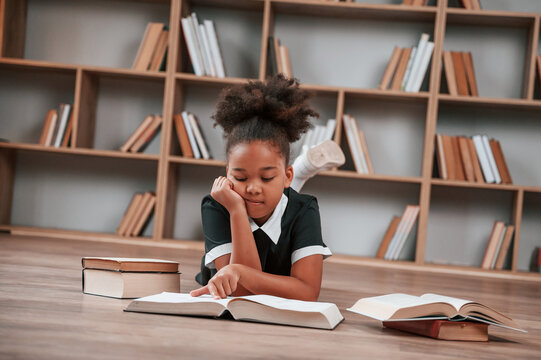 Sitting by the table with books. Cute african american girl in school uniform is at home library