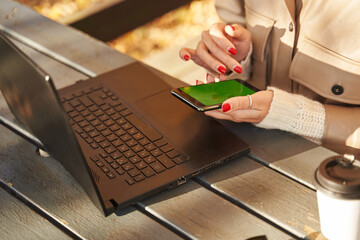 open laptop on the table and a smartphone with a green screen in female hands. there is a paper cup with coffee next to it