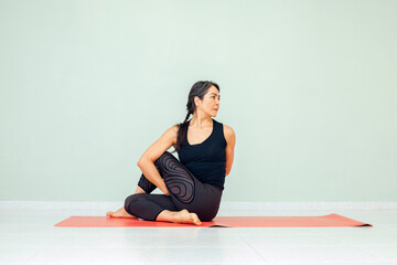 Latin woman with braid in Ardha Matsyendrasana exercise, deepened variation of Half lord of the fishes yoga pose. Working out, wearing black sportswear. Indoor full length, studio background..