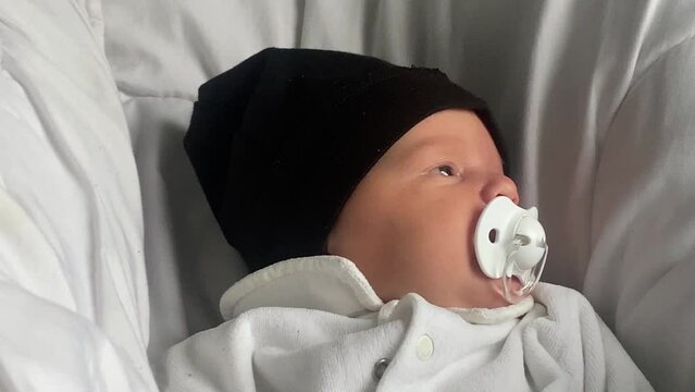 Face of a newborn baby in a black cap and pacifier on a white bed.