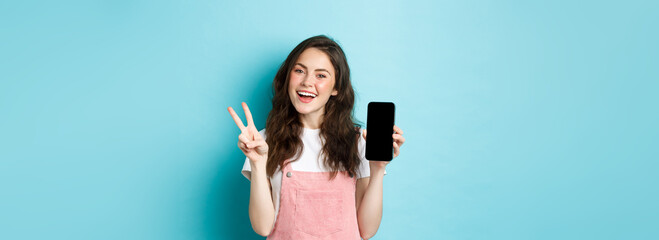 Cute young woman smiling and showing v-sign with empty smartphone screen, demonstrate app or mobile...