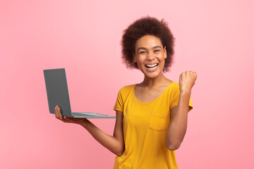 Cheerful excited young mixed race woman with curly hair rejoices online success with laptop, making...