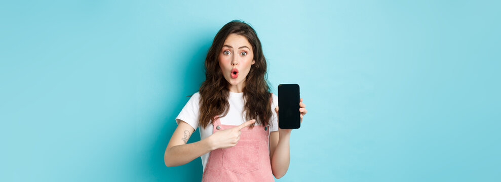 Impressed Young Modern Woman Pointing At Smartphone Screen, Say Wow And Stare Amazed At Camera, Showing Awesome Application, New Online Store, Standing Over Blue Background