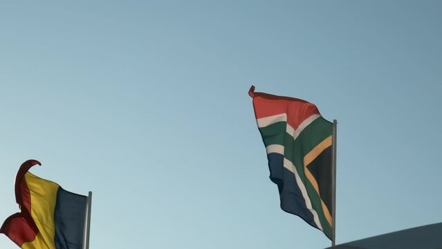 Slow motion footage of waving South Africa flag and Romania flag on a blue sky background.