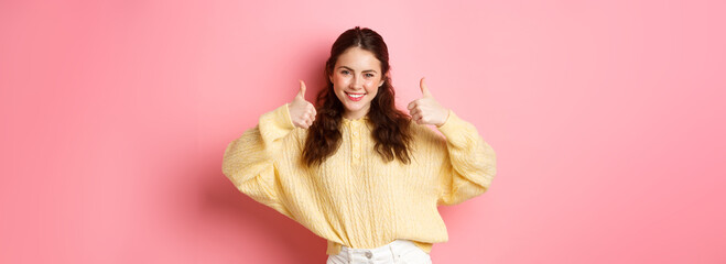 Well done, very good. Smiling young woman showing thumbs up in approval, like your choice, praise good thing, standing satisfied against pink background - obrazy, fototapety, plakaty