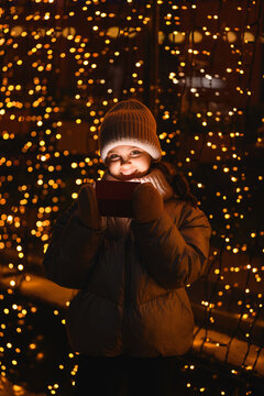 Girl and gift box with Christmas lights. Smiling girl in a hat, scarf looking at the camera at night with blurry lights. Holidays theme