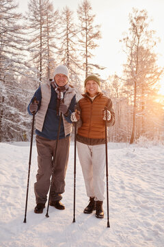 Vertical portrait of active senior couple enjoying Nordic walk with poles in winter forest and looking at camera