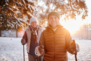 Waist up portrait of smiling mature couple enjoying Nordic walk in winter forest at sunset - Powered by Adobe