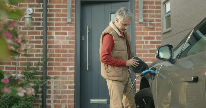 Mid Adult Male Charging Electric Car at Home and Walking Inside
