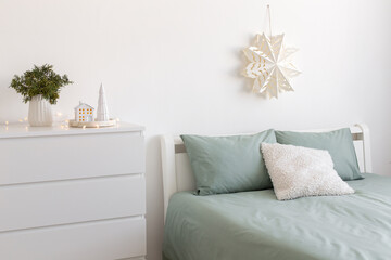Christmas home decor. Winter bedroom decor. New year interior decorations. White paper snowflake on wall, bed with green linen, fir branch in vase, decorative ceramic house, glowing garland lights.