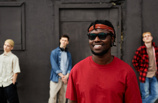 Diverse Group Of Boys Wearing Street Style Clothes Standing Against Black Wall Outdoors And Looking At Camera, Focus On Smiling African American Man