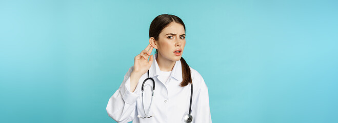 Image of confused woman doctor cant hear you, holding hand near ear and looking puzzled, speak...