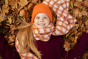 Portrait of smiling school aged girl in cute warm hat, scarf and cozy knitted sweater lying on yellow dry leaves, top view. Happy child in stylish autumn outfit walk and relax in golden park
