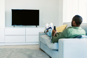 Black man watching TV with blank screen, holding remote control and switching channels on...