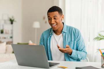 Fototapeta premium Smiling millennial black guy in jeans with wireless headphones has video call in room office interior