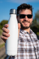 Hombre apuesto y sonriente mostrando botella metálica en blanco, para anuncios o propagandas. Fotografía con enfoque selectivo en la botella