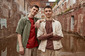 Neutral waist up portrait of two twin guys looking at camera in shabby urban setting with brick walls