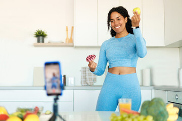 Black female fitness coach recording video on cellphone, showing donut and apple, telling about healthy and junk food