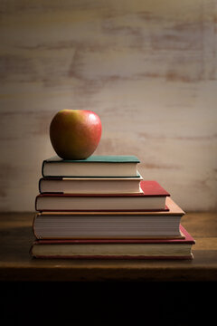 Vertical image of apple atop stack of colorful books on wooden desk against textured white backdrop
