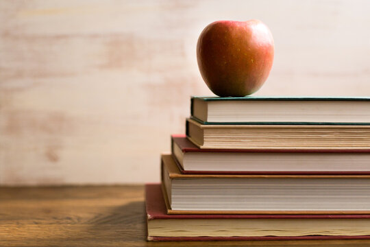 Shiny red apple sitting atop a stock of colorful books in front of a white textured background with room for copy