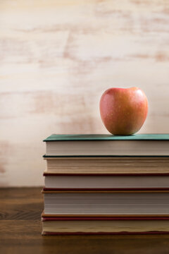 Vertical image of a shiny red apple sitting atop a stock of colorful books in front of a white textured background with room for copy