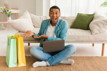 Glad excited young black man with laptop shows a lot of packages with purchases, sits on floor in...