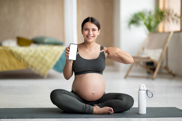 Fototapeta na wymiar Fitness App. Pregnant Woman Sitting On Yoga Mat And Showing Blank Smartphone