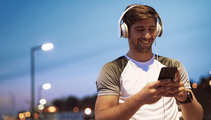 Young sporty man in active wear listening to music on big white headphones, walking the city, happy after the workout - 541335106