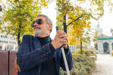 Blind bearded grey-haired mature man with dark eyeglasses on and a suit jacket holding a walking...