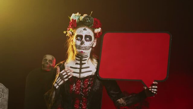 Female model showing isolated speech bubble cardboard, wearing santa muerte costume and skull make up. Person using blank copyspace template on carton board, holy goddess of death. Handheld shot.