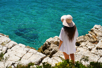 Attractive woman with a summer straw hat standing on white rocks looking at the beautiful turquoise sea
