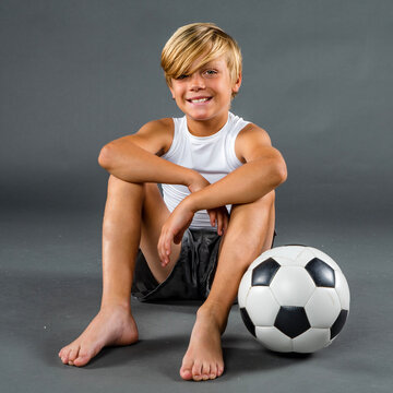 Happy young soccer player sitting barefoot on the ground with his soccer ball