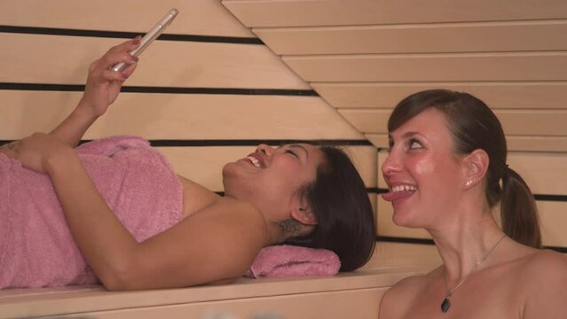 CLOSE UP: Cheerful young ladies taking selfies while having a wellness treatment. Beautiful women wrapped in pink towel using smartphone in Finnish sauna to create photos for social media content.