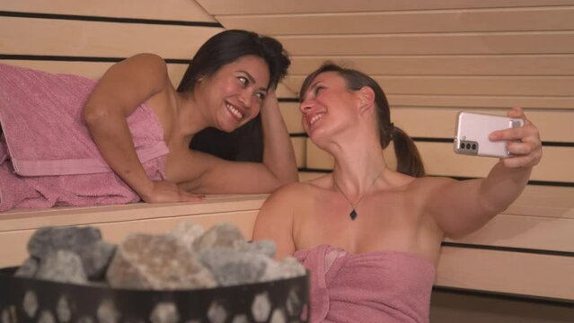 CLOSE UP: Smiling young women taking selfies with smartphone in finish sauna. Beautiful girlfriends wrapped in pink towel having a wellness treatment and creating photos for social media content.