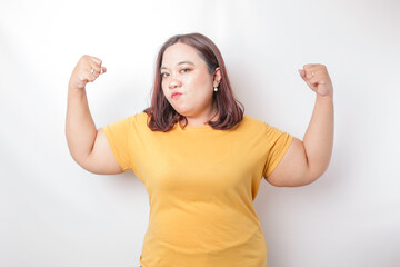 Excited Asian big sized woman wearing a yellow t-shirt showing strong gesture by lifting her arms...