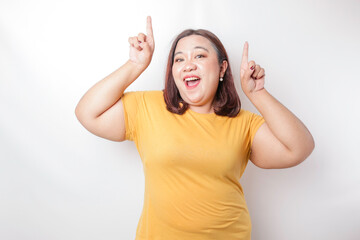 Excited Asian big sized woman wearing yellow shirt pointing at the copy space upside her, isolated...