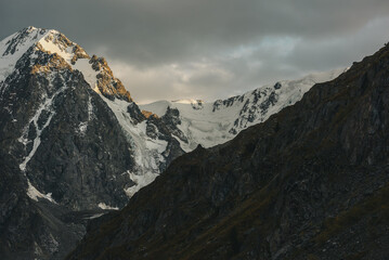 Gloomy landscape with giant snowy mountain range and sunlit gold rocks in dramatic gray sky. Huge...