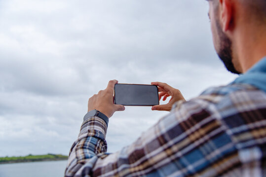 Hombre joven de perfil con camisa de cuadros y un reloj inteligente sacando una foto al paisaje  con dos manos con su teléfono móvil en horizontal durante un día nublado en la playa de El Sardinero