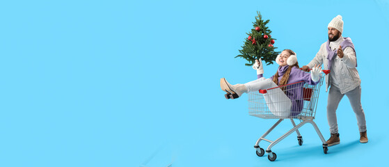 Happy young couple with Christmas tree and shopping cart on light blue background with space for text