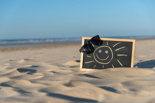 Pizarra negra con marco de madera con un sol sonriendo dibujado con tizas de colores y unas gafas de sol en una esquina sobre la arena dorada de la playa y con el mar de fondo durante un d&iacute;a soleado